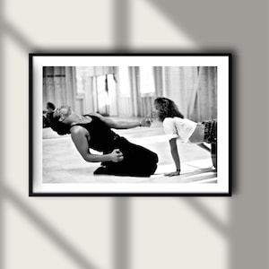 May include: A black and white framed photograph depicting two individuals in a dance studio. One individual is kneeling and leaning backward in a tank top and trousers, while the other is positioned on the floor in a white t-shirt and shorts.