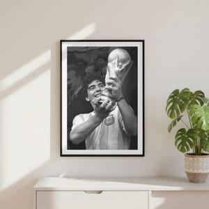 May include: A black and white framed photograph depicting a football player wearing an AFA jersey, smiling while holding the FIFA World Cup trophy high above their head with both hands. The image captures a celebratory moment in a stadium setting.