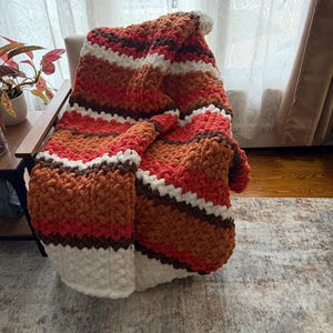 May include: A chunky knit throw blanket featuring horizontal stripes in shades of burnt orange, deep red, dark brown, and cream. The blanket is crafted from thick, soft yarn with a visible looped texture, creating a dense and cosy appearance.