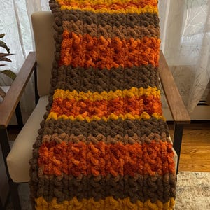 May include: A chunky knit throw blanket featuring horizontal stripes in shades of mustard yellow, burnt orange, and dark brown. The blanket is crafted from thick, soft yarn loops, creating a textured, voluminous appearance with a consistent pattern.