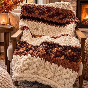May include: A chunky knit throw blanket featuring horizontal stripes in cream, burnt orange, dark brown, and variegated autumn-toned yarn. The blanket has a thick, textured loop construction and is draped over a wooden armchair with light upholstery.