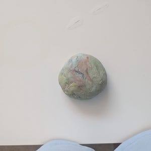 Può includere: Un oggetto arrotondato simile a una pietra con una superficie ruvida caratterizzata da una tavolozza di colori screziati di verde salvia pallido, rosa tenue e sottili vortici grigio-blu. L'oggetto ha una forma organica e irregolare.
