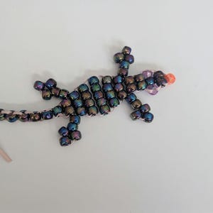 May include: A lizard-shaped ornament crafted from iridescent dark blue and purple metallic beads. The figure features a long tail, four legs, and a head accented with two light purple beads for eyes and a single bright orange bead at the snout.