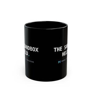May include: A black ceramic mug featuring white text that reads THE SANDBOX HELD. Below the text, smaller white lettering says MODEL & MACHINE, accompanied by a small light blue square icon and a light blue letter M logo on the side.
