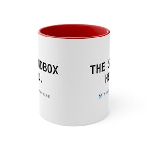Op de afbeelding: Een witte keramische mok met een felrode binnenkant. De buitenkant is voorzien van zwarte tekst met de tekst THE SANDBOX HEAD en MODEL MACHINE, samen met een klein blauw logo. Het ontwerp is gedrukt op het gladde witte oppervlak.