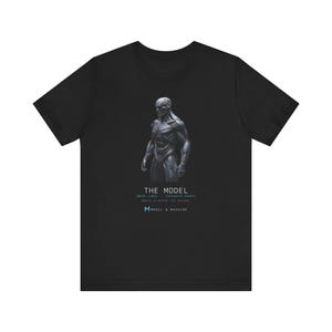 May include: A black short-sleeved t-shirt featuring a graphic of a grey, muscular, robotic humanoid figure. Below the figure, the text reads THE MODEL, followed by smaller text and the blue letter M with the words MODEL & MACHINE.