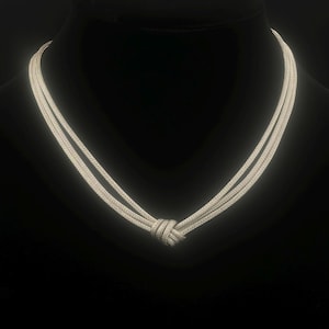 May include: A silver-toned multi-strand necklace featuring a central knot detail. The piece consists of several thin, textured metallic chains that converge into a single, tightly wound knot at the bottom center, creating a layered appearance.