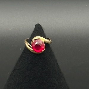 May include: A gold ring featuring a single, round, faceted red gemstone set in a bypass-style band. The metal band curves around the central stone, creating a smooth, continuous design that highlights the vibrant color and clarity of the red gem.