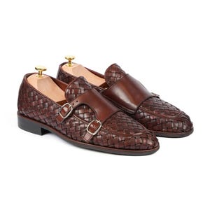 Pode incluir: Um par de sapatos de fivela dupla castanho-escuro com parte superior em pele entrançada, uma tira de pele lisa sobre o peito do pé e duas fivelas metálicas prateadas. Cada sapato inclui uma forma de madeira clara com um botão dourado.