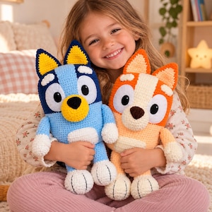 Op de afbeelding: Twee handgemaakte gehaakte knuffels die tekenfilmhonden voorstellen. De ene is lichtblauw met donkerblauwe vlekken en gele oren, de andere is oranje met crèmekleurige vlekken en oranje oren. Beide hebben witte snuiten en zwarte neuzen.