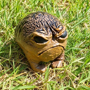 May include: A small, round, brown creature figurine with a textured, bumpy surface, deep-set black eyes, a furrowed brow, and a downturned mouth. The figure has short, stubby limbs and a wide, squat body shape, crafted with detailed skin folds.