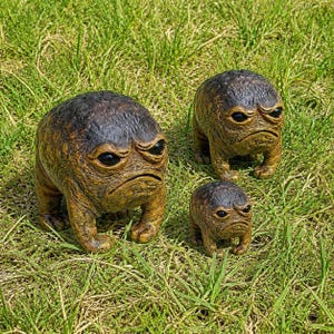May include: Three brown, textured, four-legged creature figurines with dark, glossy eyes and downturned mouths. The figures have a rounded, bumpy body shape and are arranged in a group, with two larger ones and one smaller one, all featuring a grumpy expression.