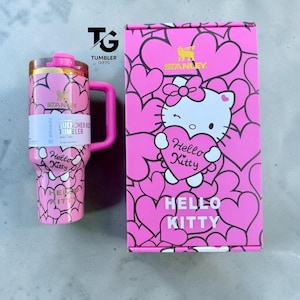 May include: A pink Stanley Quencher H2.0 tumbler featuring a Hello Kitty heart pattern and a matching pink box with a Hello Kitty graphic, heart motifs, and the text Hello Kitty and Stanley. The tumbler includes a handle and a pink lid.