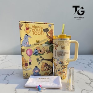Puede incluir: Un vaso Stanley Quencher H2.0 amarillo con personajes de Winnie the Pooh, una caja a juego con ilustraciones, un paño blanco con el logo de Stanley, una pajita transparente en una funda y un pequeño dije de personaje azul.