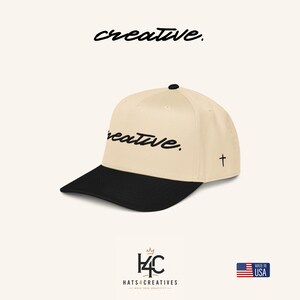 Pode incluir: Um boné de beisebol bege com aba preta e um botão preto no topo. A frente apresenta a palavra creative em letra cursiva preta. Uma pequena cruz preta está bordada na lateral. A palavra creative também está impressa acima do boné.