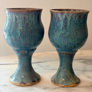 May include: Two ceramic goblets featuring a bulbous bowl and a flared base. The glaze displays a mottled blend of deep teal, blue, and brown tones with a glossy finish. Each goblet has a small, circular stamped mark on the stem near the bowl.