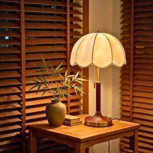 Op de afbeelding: Een tafellamp met een crèmekleurige, geschulpte stoffen kap met een subtiel bloemmotief, een cilindrische standaard van donker hout en een messing voet. Een trekketting hangt aan de kap en de lamp geeft een warm, zacht licht.