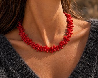 Puede incluir: Un collar compuesto por numerosos segmentos pequeños, irregulares y de color rojo brillante con forma de rama. Las piezas están unidas para formar un collar denso y texturizado que se asienta cerca de la base del cuello, creando un contraste vibrante con el suéter gris oscuro.