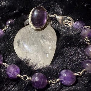 May include: A silver ring featuring an oval purple amethyst stone with a twisted metal border, accompanied by a matching bracelet composed of faceted purple amethyst beads linked together with silver wire, resting near a clear heart-shaped stone.