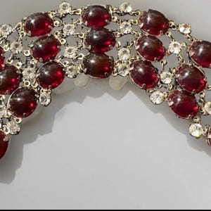 May include: A wide bracelet featuring three rows of oval-shaped, deep red cabochon stones set in a gold-tone metal frame. The design is accented with small, clear, round faceted crystals interspersed between the red stones and includes a circular clasp.