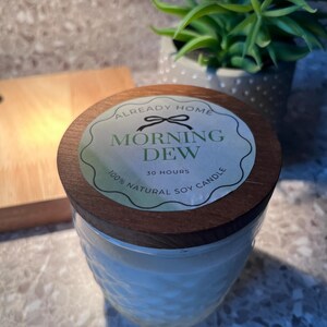 May include: A glass jar candle with a wooden lid. The label reads "Morning Dew" and "100% Natural Soy Candle". The candle has a textured pattern and is next to a small wooden cutting board and a potted plant.