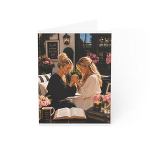 May include: A greeting card featuring two individuals with blonde hair, one in a black top and one in a white dress, holding hands over an open book. A mug labeled Faith Over Fear, a candle labeled Grace, and books titled Streams in the Desert and Gentle and Lowly are present.