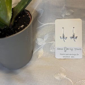 Op de afbeelding: Een paar zilverkleurige libelle-oorbellen op een witte displaykaart. De kaart bevat de tekst Sens-Ear-ly Yours en Plastic post earrings for sensitive ears. De oorbellen hebben doorzichtige plastic haakjes en kleine metalen bedels.
