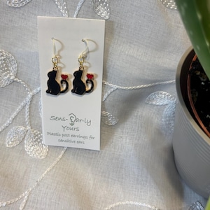 May include: A pair of dangle earrings featuring black cat silhouettes with small red hearts attached to their tails. The earrings are displayed on a white card labeled Sens-Ear-ly Yours, which notes plastic posts for sensitive ears.