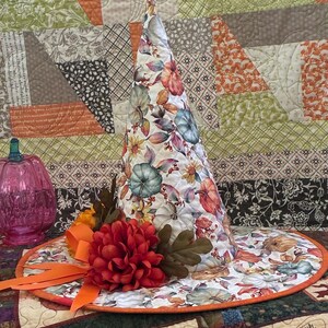 May include: A quilted fabric witch hat featuring a white background with a colorful autumn print of pumpkins, leaves, and flowers. The hat is accented with a large red fabric flower, brown fabric leaves, and an orange ribbon bow on the brim.