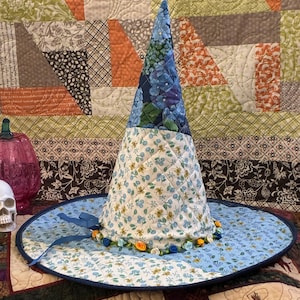 May include: A quilted fabric hat with a tall, pointed crown and a wide brim. The crown features a blue floral pattern on top and a white base with small yellow and blue flowers. The brim is light blue with a dark blue edge and small fabric roses.