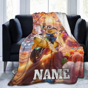 May include: A plush throw blanket featuring a colourful graphic of yellow Minion characters riding banana-shaped scooters on a candy-themed path. The design includes vibrant orange, yellow, and pink hues with the word NAME printed in large white letters.