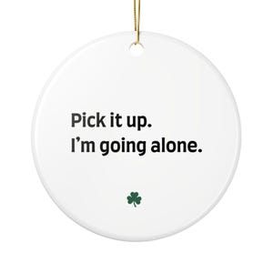 Puede incluir: Un adorno redondo de cerámica blanca con el texto negro Pick it up. I’m going alone. centrado en la parte frontal. Debajo del texto hay un pequeño icono de trébol verde. El adorno incluye un cordón dorado en la parte superior para colgar.