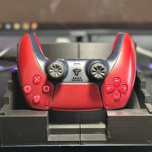 May include: A metallic red and dark gray video game controller resting in a black plastic stand. The controller features two analog sticks, a directional pad, four action buttons with geometric symbols, and a central touchpad with a logo.