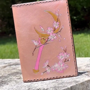 May include: A light tan leather passport holder featuring a debossed and painted graphic of a golden crescent moon wand with pink cherry blossoms. The item has visible dark brown stitching along the edges and a smooth, rectangular surface.
