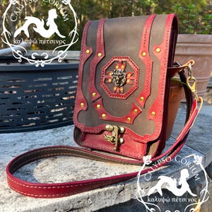 May include: A brown and burgundy leather crossbody bag with gold-colored accents. The bag features a decorative front panel with a Medusa head emblem and a clasp closure. The bag has a long, burgundy strap.