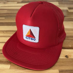 May include: A bright red trucker hat featuring a structured front panel and a breathable mesh back. A square white patch is centered on the front, displaying a colorful triangular logo in orange, red, and dark brown with the text CITGO in blue.