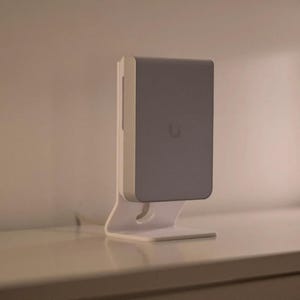 May include: A rectangular wireless access point with a light gray front panel and a white chassis, mounted on a white plastic stand. The device features a small, centered logo on the front face and is positioned vertically on a flat surface.
