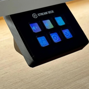 Pode incluir: Um controlador Stream Deck preto com seis teclas quadradas e ícones azuis iluminados. O dispositivo está montado num ângulo e apresenta símbolos para silenciar áudio, uma função de pausa, um rosto sorridente e captura de ecrã.