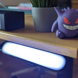 May include: A purple Gengar figurine with red eyes and a wide grin, positioned on a wooden desk next to a gray potted plant and a black rectangular speaker, with a horizontal white LED light bar mounted underneath the desk edge.