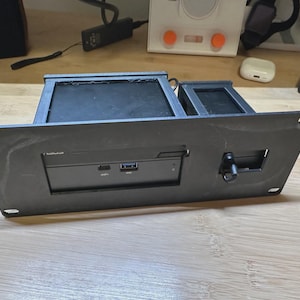 May include: A black 3D-printed mounting bracket featuring a rectangular faceplate with four corner screw holes. The center cutout houses a small electronic device with a USB-C port, a USB-A port, and a small black toggle switch on the right side.