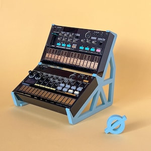 May include: A light blue angled stand holding two black Korg Volca synthesizers, the Volca FM and Volca Beats, stacked vertically. A small, light blue circular symbol with a diagonal line through it sits on the surface next to the stand.
