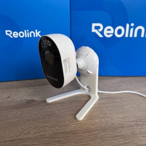 May include: A white Reolink security camera with a black front faceplate featuring a lens and sensor. The camera is mounted on a white adjustable stand and includes a white power cable extending from the back. The Reolink logo is visible on the camera body.