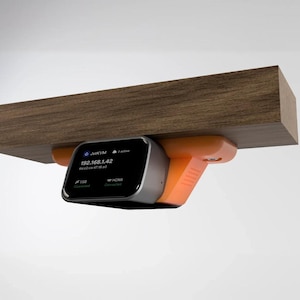 May include: A JatKVM device with a black screen displaying 192.168.1.42, USB, and HDMI status. The device is housed in a silver and orange casing and is mounted to the underside of a rectangular wooden shelf with a visible wood grain pattern.