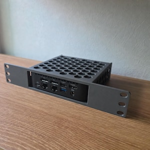 May include: A dark gray metal rack mount bracket holding a small black computer device. The bracket features a hexagonal honeycomb pattern on the top surface and includes side flanges with circular mounting holes for installation.