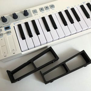 May include: A white Arturia KeyStep MIDI keyboard controller with black and white keys, control knobs, and touch strips, positioned above two black, rectangular, open-frame stands with a hollow center design and small mounting points.