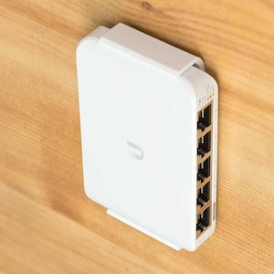 May include: A white network switch mounted on a wall, featuring a rectangular design with rounded corners, a central logo, and five Ethernet ports on the side. The device includes small indicator lights near the top of the port panel.