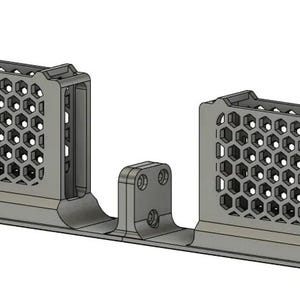 Puede incluir: Un soporte de montaje gris y alargado que presenta dos torres rectangulares verticales con una malla de patrón de panal distintiva en los lados. La base es plana con pequeños orificios circulares en cada extremo y una pestaña central con tres orificios.