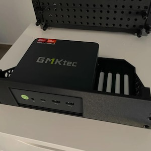 Può includere: Un mini computer GMKtec nero montato in una staffa per rack nera personalizzata. Il pannello frontale presenta un pulsante di accensione verde, due porte USB e una porta USB-C. La superficie superiore mostra il logo GMKtec e adesivi rossi AMD Radeon e Ryzen.