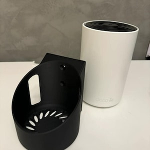 May include: A white cylindrical Deco BE mesh Wi-Fi unit with a black top and a black plastic wall mount bracket featuring a circular base, side ventilation slots, and two mounting holes on the back panel for secure installation.