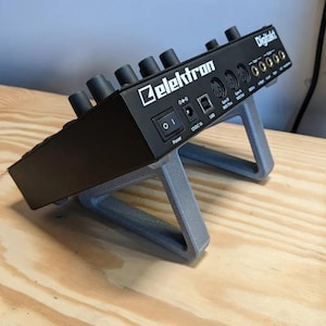Op de afbeelding: Een zwarte Elektron Digitakt drumcomputer en sampler op een grijze schuine standaard. Het achterpaneel bevat een aan-uitknop, een 12VDC 1A stroomingang, een USB-poort, MIDI In, Out en Thru poorten, en audio in- en uitgangsaansluitingen.